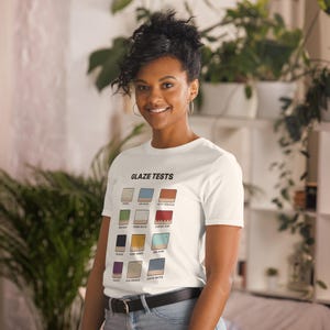 May include: A white short-sleeve t-shirt featuring the text GLAZE TESTS at the top, followed by a grid of twelve colorful square icons representing different glaze samples, each labeled with its specific color name in black text.