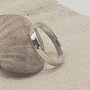 May include: A simple silver band ring with a smooth, rounded surface. The ring is on a light brown fabric background.