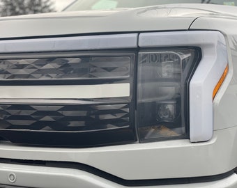 May include: Close-up of a white pickup truck's front grille and headlights. The grille is black with a diamond pattern and a white horizontal stripe. The headlights are black with a clear lens and a white accent.