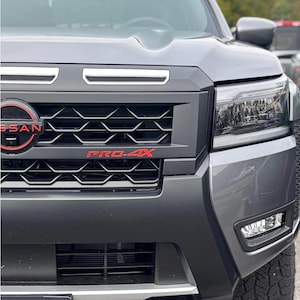 Peut inclure: Gros plan sur l'avant d'un camion Nissan gris. La calandre affiche le logo Nissan en rouge et le texte "PRO-4X" en rouge. Le camion a des détails noirs, des phares et un crochet de remorquage rouge.