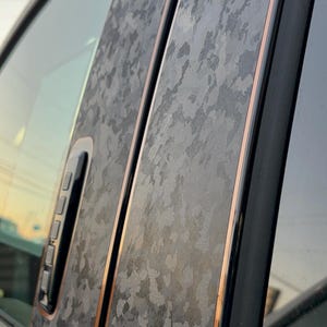 Könnte beinhalten: Nahaufnahme einer Autotür mit einer schwarzen und grauen Tarnmuster-Vinylfolie. Die Folie hat einen kupferfarbenen Rand an den Kanten.