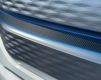 May include: Close-up of a car's front end, showcasing a silver grille with a diamond pattern. A carbon fiber-style accent is framed by a bright blue trim. The hood features a geometric design in shades of gray.