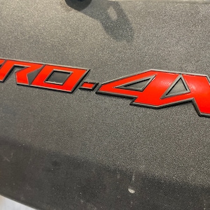 May include: A red "PRO-4X" emblem with black outlining. The emblem is on a black textured surface.