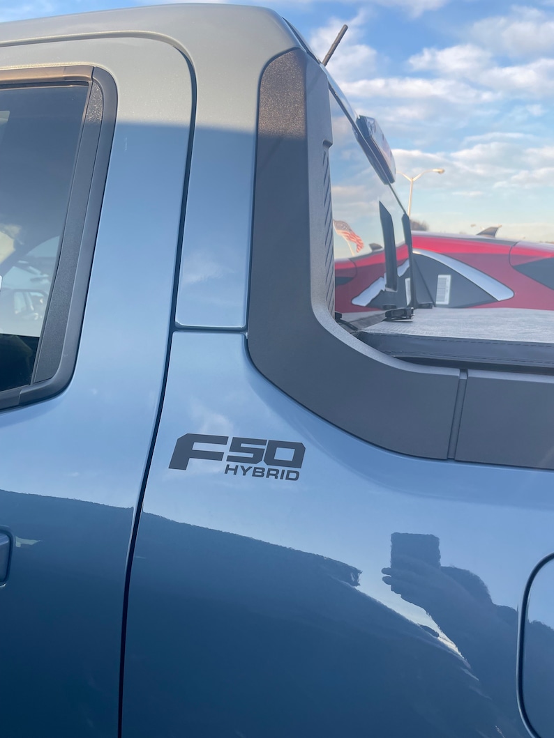 May include: A light blue pickup truck with the black text "F50 HYBRID" on the side. The truck has a black trim around the windows and a black antenna. The sky is visible in the background.