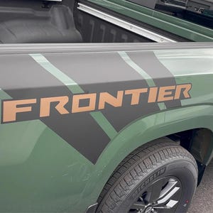 May include: A green pickup truck with a black and green graphic on the side of the truck bed. The graphic includes the word "FRONTIER" in a copper color.