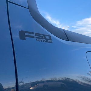 May include: Close-up of a blue vehicle's side panel with the black text "F50 HYBRID". The vehicle's surface reflects the sky and surrounding environment. The image is taken outdoors.