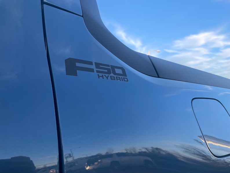 May include: Close-up of a blue vehicle's side panel with the black text "F50 HYBRID". The vehicle's surface reflects the sky and surrounding environment. The image is taken outdoors.
