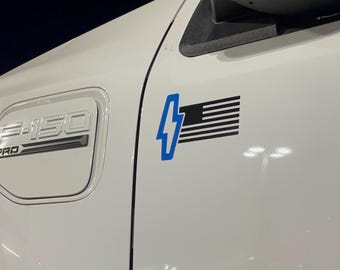 May include: White F-150 Pro electric truck with a blue lightning bolt and American flag graphic. The truck's charging port door is visible, along with the "F-150 PRO" emblem.