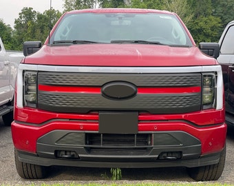 May include: A red Ford F-150 Lightning electric pickup truck with a black grille and a red accent stripe. The truck has a black bumper and black headlights.