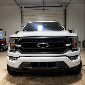 Peut inclure: Un camion Ford F-150 blanc avec une calandre noire et un logo Ford ovale chromé.