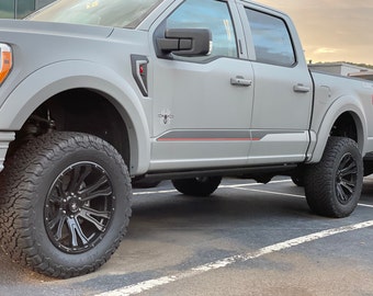 Może przedstawiać: Srebrny samochód ciężarowy Ford F-150 z czarnymi kołami i oponami. Samochód ma czarny i czerwony pasek po bokach. Samochód jest zaparkowany na parkingu.
