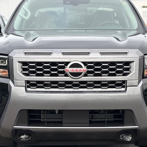 May include: Close-up of a black and silver Nissan truck front. The grille features a honeycomb pattern with the Nissan logo in the center. Headlights and fog lights are visible. The vehicle is parked outdoors.