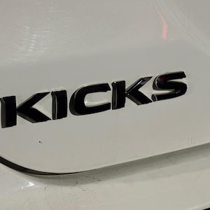 May include: A close-up shot of a white car panel featuring the word "KICKS" in black, glossy lettering. The letters are in a bold, sans-serif font and appear to be raised. The car's surface reflects light.