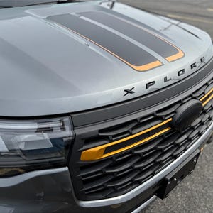 May include: Close-up of a gray vehicle's front end. The hood features black and orange stripes. The word "XPLORE" is displayed above the black and orange grill. The grill has orange accents.