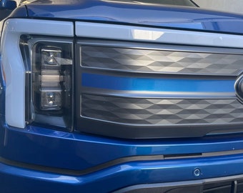 May include: A blue pickup truck with a black and gray diamond-patterned grille. The grille has a blue stripe across the center. The truck has a white stripe along the top of the hood.