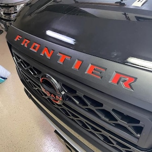 May include: A black Nissan Frontier pickup truck with red "FRONTIER" lettering on the front grille. The truck has a black grille with a honeycomb pattern and a Nissan logo in the centre.