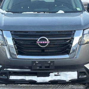 Peut inclure: Vue de face d'un SUV Nissan gris. Le véhicule présente une calandre noire avec une bordure chromée et le logo Nissan au centre. Le pare-chocs avant est noir et les phares sont visibles de chaque côté.