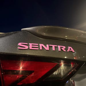 Könnte beinhalten: Ein dunkelgrauer Kofferraumdeckel mit dem Wort SENTRA in fetten, leuchtend rosa Buchstaben. Das Fahrzeug verfügt über einen Heckspoiler über dem Emblem sowie eine rote und klare Rückleuchteneinheit am unteren Bildrand.
