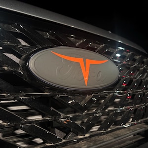 May include: A black vehicle grille featuring a matte gray oval emblem centered in the middle. The emblem displays a bright orange stylized T-shaped graphic overlaying a faint, embossed script logo. The grille has a glossy black geometric pattern.