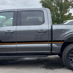Könnte beinhalten: Ein grauer Pickup mit einem dekorativen Vinyl-Aufkleberset an den Seitenwänden. Das Dekor besteht aus schwarzen horizontalen Streifen mit orangefarbenen Akzentlinien, und das Wort COYOTE ist in Orange auf dem hinteren Seitenteil aufgedruckt.