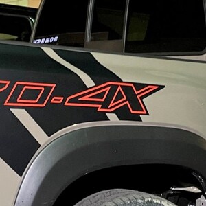 Puede incluir: Primer plano de una camioneta gris con una calcoman&iacute;a de rayas negras y blancas y un emblema rojo "PRO-4X" en el lateral.