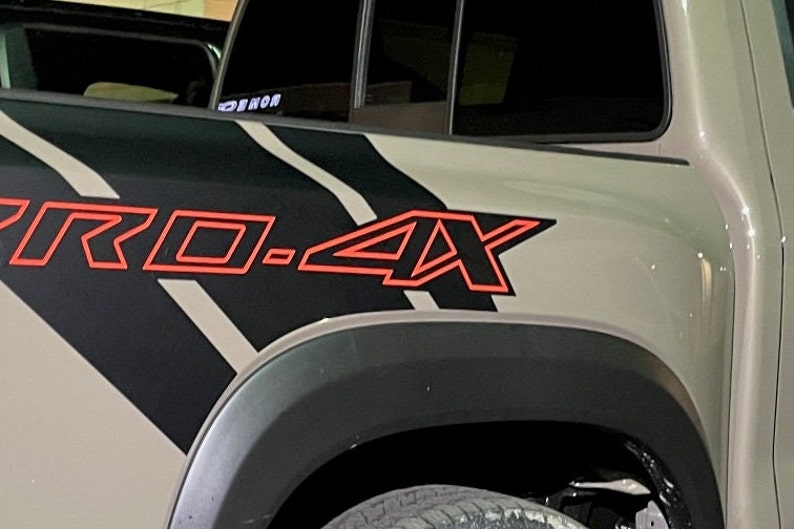 Puede incluir: Primer plano de una camioneta gris con una calcoman&iacute;a de rayas negras y blancas y un emblema rojo "PRO-4X" en el lateral.