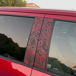 Op de afbeelding: Rode pick-up truck met zwarte raambekleding en rode en zwarte gepatroneerde raamstickers. De stickers hebben een topografisch kaartontwerp.