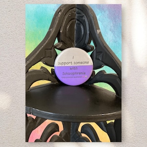 May include: A round button with the text "I support someone with Schizophrenia" in white and purple. The button is resting on a black decorative shelf with a colorful watercolor background.