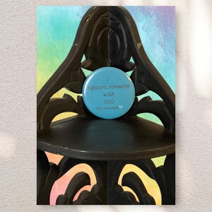 May include: A light blue button with the text "I support someone with OCD" and "OCD Awareness" on a black decorative shelf. The background features a watercolor wash of blue, green, and purple.