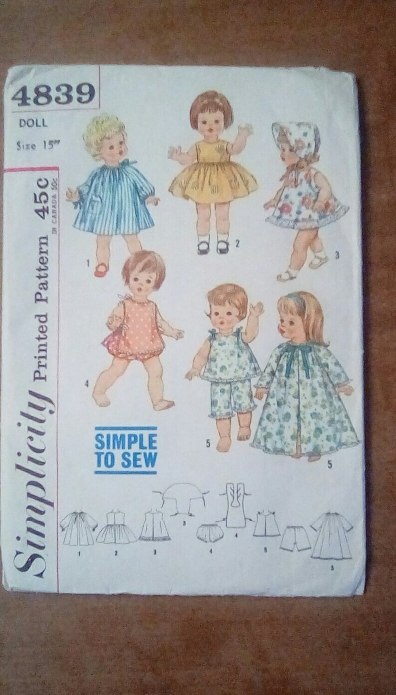 May include: A vintage Simplicity sewing pattern for a 15-inch doll. The pattern includes five different outfits: a blue and white striped dress, a yellow dress, a floral print dress, a floral print pajamas set, and a floral print robe. The pattern is labeled "Simplicity Printed Pattern 45c" and "Simple to Sew".