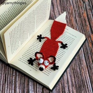 May include: A crocheted fox bookmark in red, white, and black. The fox has googly eyes and a white tipped tail. The bookmark is placed in an open book.