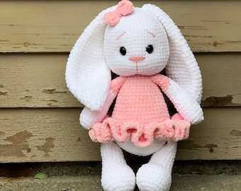 May include: A white crocheted bunny wearing a pink dress with a white bow on its head. The bunny is standing in front of a wooden wall.