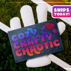 May include: A blue denim card with the text "Cozy Crafty Chaotic" in rainbow gradient letters. The card is held by a white hand with the text "Ships Today!" in a pink and purple box above the hand.