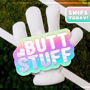 May include: A white hand with a rainbow sticker that says "BUTT STUFF" on it. The sticker has a heart shape on the left side. The background is green.