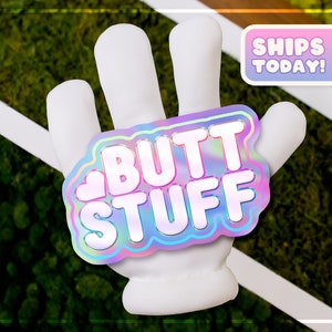 May include: A white hand with a holographic sticker that says "BUTT STUFF" with a pink heart in the corner. The sticker has a rainbow iridescent effect. The background is green moss.