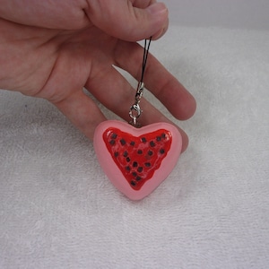 May include: A pink heart-shaped charm with a red watermelon slice design and black seeds. The charm is attached to a black string.