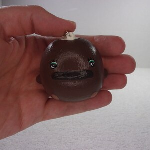 May include: A small brown ceramic ornament with a painted face. The ornament has green eyes, a black mouth, and a white top.