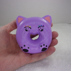 May include: Purple donut shaped squish toy with cat ears, painted black paw prints and rainbow colored eyes.