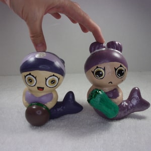 以下が含まれることがあります： 紫色の髪、大きな目、薄い茶色の肌をした、手描きのセラミック製人魚像2体。1体の人魚は緑色の海藻のようなものを持ち、もう1体の人魚は茶色のものを持ちます。