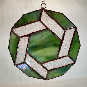 Puede incluir: Un atrapasol de vidrio verde y blanco con un diseño geométrico. El atrapasol está hecho de múltiples piezas de vidrio que se unen con lámina de cobre. El atrapasol está colgado de una cadena de cobre.