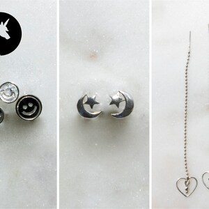 May include: Three pairs of silver earrings. The first pair features a smiley face design. The second pair features a crescent moon and star design. The third pair features a long chain with a heart charm at the end.