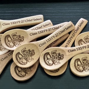 May include: A collection of light-colored wooden spoons, each featuring a laser-engraved USN anchor emblem on the bowl and the text Class 133 FY-27 Chief Select printed on the handle, arranged in a scattered pile.