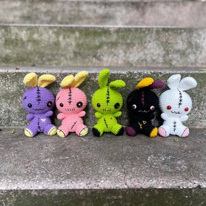 May include: Five crocheted plush bunny toys with stitched-on eyes and mouths. The bunnies are purple, pink, green, black, and white. They are sitting on a set of concrete steps.