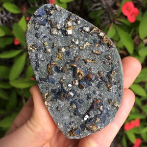 May include: A gray, irregularly shaped stone with embedded pyrite crystals. The stone has patches of gold, blue, and rust-colored minerals. The surface is textured, and the overall appearance is natural and earthy. The stone is held in a hand.