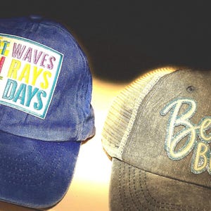 Pode incluir: Dois bonés de beisebol. Um é azul com o texto "BOAT WAVES SUN RAYS LAKE DAYS" em um patch colorido. O outro é cinza com uma parte traseira de malha e o texto "Beach Bum" em itálico.