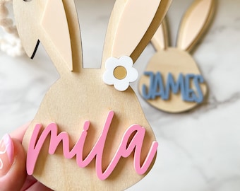 Könnte beinhalten: Osterhasen-Kopfschmuck aus Holz mit geschichteten Ohren und einem Gänseblümchen-Akzent. Der Name "mila" ist in rosa Schrift geschrieben. Ein zweiter Hasenschmuck mit dem Namen "james" ist im Hintergrund zu sehen.