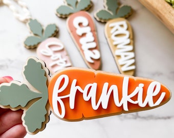 May include: Wooden carrot-shaped name tags with white lettering. The tags are orange, pink, and tan, with green leaf accents. Names include Frankie, Cruz, Roman, and other names. The tags are likely for Easter or spring decor.