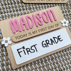 May include: A wooden sign featuring the name MADISON in raised pink letters, with the text TODAY IS MY FIRST DAY OF and a white rectangular insert displaying the handwritten text FIRST GRADE, accented by two white and gold flower cutouts.
