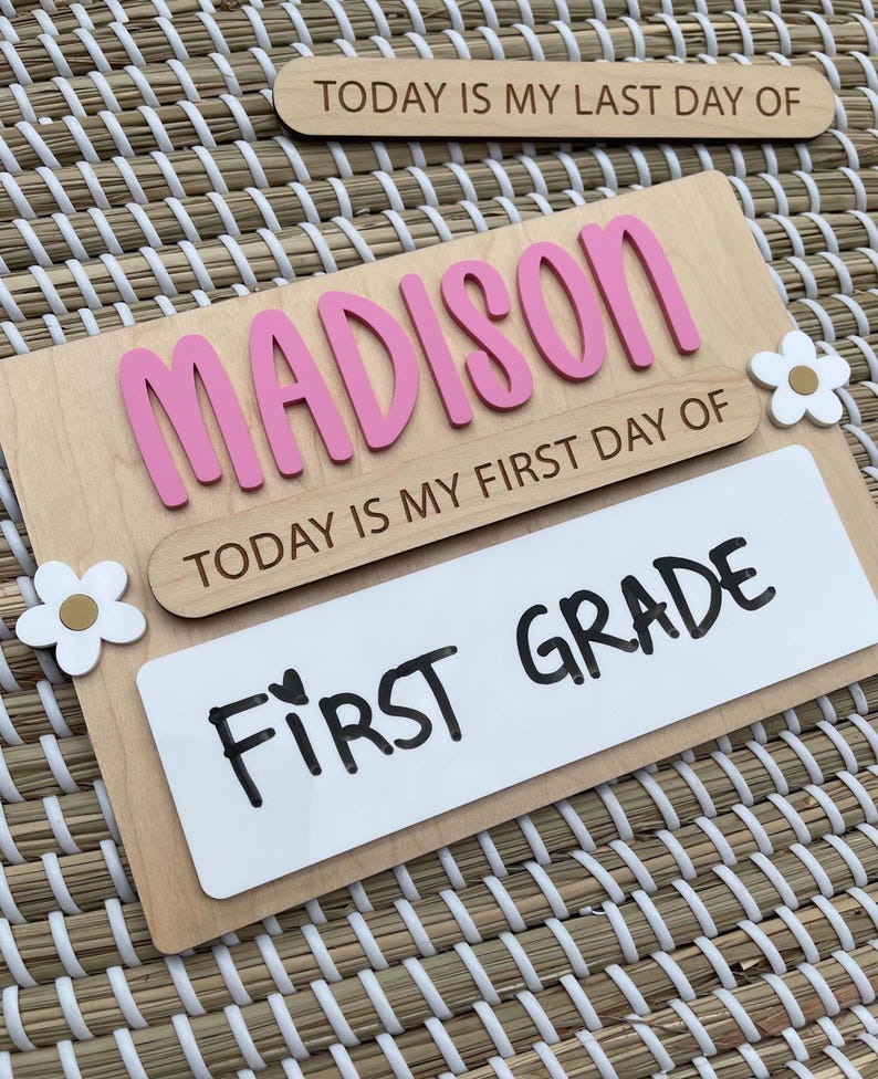 May include: A wooden sign featuring the name MADISON in raised pink letters, with the text TODAY IS MY FIRST DAY OF and a white rectangular insert displaying the handwritten text FIRST GRADE, accented by two white and gold flower cutouts.
