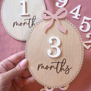 May include: Wooden baby milestone markers with pink bows and white numbers. The markers are in the shape of a circle and have a brown background. The numbers are 1, 3, 4, 5, 6, 8, and 9. The text on the markers reads "1 month" and "3 months".